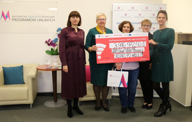 Pięć osób stojących, trzy z nich trzymają tablice informacyjną z podpisania umowy projektu pn. Sięgnij po więcej, dofinansowanego z RPOWM 2014-2020.